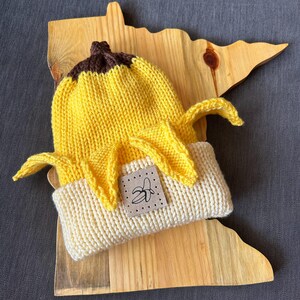 May include: A knitted beanie shaped like a peeled banana, featuring a bright yellow body, a dark brown top, and a cream-colored folded brim. A square beige patch is stitched to the front, displaying a simple black line drawing of a banana.
