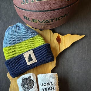 May include: A blue, green, and navy blue knit beanie with a leather patch featuring a silhouette of a wolf. Two white knit baby booties with a wolf graphic and the text "HOWL YEAH" are also shown. A Spalding basketball is in the background.