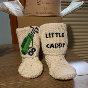 May include: White knitted baby booties with embroidered designs. One bootie has a green golf bag with clubs and the other has the text "LITTLE CADDY".