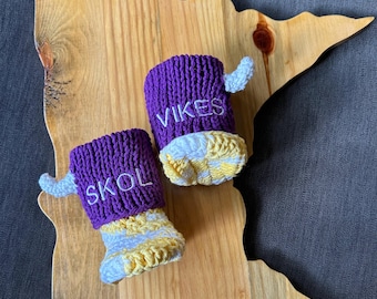 May include: Two purple and white crocheted baby booties with small white horns on the sides. The left bootie features the white embroidered text SKOL and the right bootie features the white embroidered text VIKES on the purple cuff.