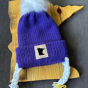 May include: A purple knit hat with a white pom-pom and white braided tassels. The hat features a square patch with a black silhouette of the state of Minnesota. The tassels are tied with yellow yarn. The hat is displayed on a wooden cutout of the state of Minnesota.