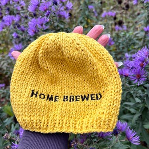 May include: A mustard yellow knit beanie featuring the text HOME BREWED embroidered in black thread across the front. The hat is constructed from a thick yarn with a visible stockinette stitch pattern and a folded brim edge.