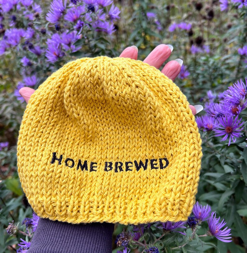 May include: A mustard yellow knit beanie featuring the text HOME BREWED embroidered in black thread across the front. The hat is constructed from a thick yarn with a visible stockinette stitch pattern and a folded brim edge.