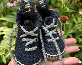May include: A pair of black crocheted baby booties designed to resemble high-top sneakers. The booties feature grey laces, a yellow stitched line along the sole, and the text LIL embroidered in yellow on the back pull tabs.