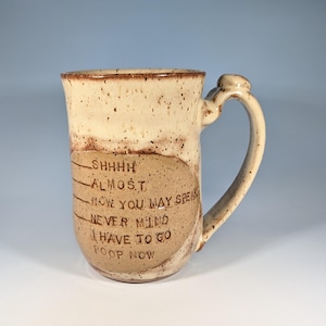 May include: A handmade ceramic mug with a speckled beige and brown glaze. The mug features a curved handle and a brown oval with etched text that reads "SHHHH, ALMOST, NOW YOU MAY SPEAK, NEVER MIND, I HAVE TO GO, POOP NOW."