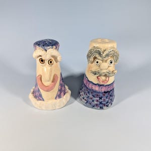 May include: A pair of whimsical ceramic salt and pepper shakers. One shaker has a blue top and a smiling face, the other has a mustache and a stern expression. Both have a cream-colored base and blue and purple patterned details.