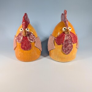 May include: Two ceramic rooster figurines. The roosters are yellow with red combs and wattles. They have pink accents around their necks and eyes. The figurines are on a white surface with a light blue background.