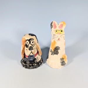 May include: Handmade ceramic dog and cat figurines. The dog has a black and tan head with a red collar and a blue tag that says "BOB". The cat is white with orange and black spots and yellow eyes. These figurines are whimsical and decorative.