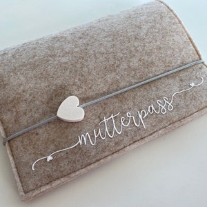 Könnte beinhalten: Eine beige Filztasche mit einem weißen Herzanhänger und dem Text "Mutterpass" in weißer Kursivschrift.