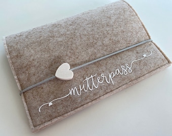 Könnte beinhalten: Eine beige Filztasche mit einem weißen Herzanhänger und dem Text "Mutterpass" in weißer Kursivschrift.