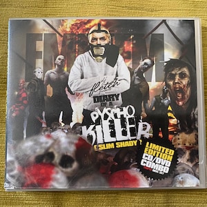 Puede incluir: Un conjunto combinado de CD/DVD con el álbum "Diary of a Psycho Killer" de Slim Shady. La portada incluye una imagen de una persona con máscara y sudadera con capucha, rodeada de figuras de zombis y el texto "Eminem".