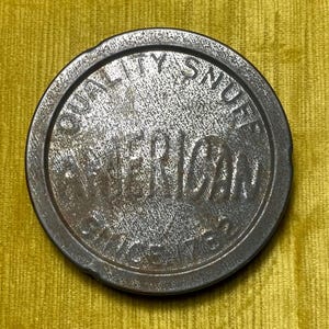 Pode incluir: Uma lata redonda de rapé metálica com as palavras "QUALITY SNUFF AMERICAN SINCE 1876" em relevo na borda. A lata tem uma aparência desgastada e envelhecida, sugerindo que é um item antigo ou vintage. O fundo é um tecido amarelo texturizado.