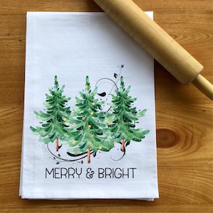 Peut inclure: Serviette de cuisine blanche avec une illustration aquarelle de trois sapins de No&euml;l avec des ornements color&eacute;s et le texte "Merry & Bright".