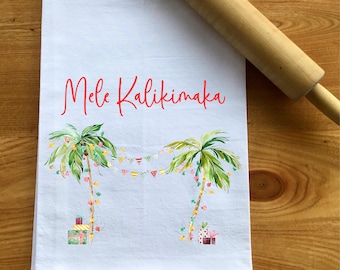 May include: White kitchen towel with a red "Mele Kalikimaka" text and a watercolor illustration of two palm trees decorated with Christmas lights and presents.