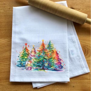 May include: A white kitchen towel with a colorful illustration of five Christmas trees decorated with ornaments and lights. The trees are arranged in a row and are surrounded by a scattering of stars.