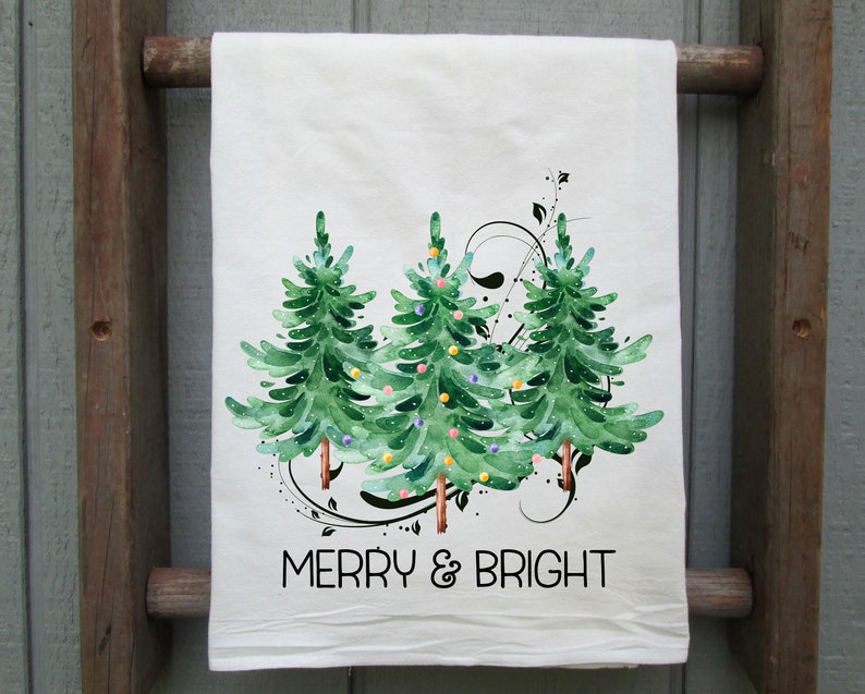 Peut inclure: Serviette de cuisine blanche avec une illustration aquarelle de trois sapins de No&euml;l verts avec des ornements color&eacute;s et le texte "Merry & Bright".