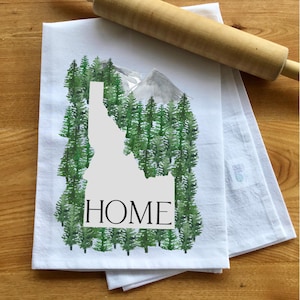 May include: White kitchen towel with a watercolor design of the state of Idaho, surrounded by green trees and a mountain range. The word "HOME" is printed in black. A wooden rolling pin rests on top of the towel.