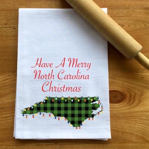 May include: White kitchen towel with a green and black plaid outline of the state of North Carolina with colorful Christmas lights. The text "Have A Merry North Carolina Christmas" is printed in red above the state.
