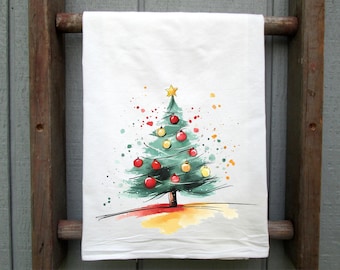 Peut inclure: Torchon de cuisine blanc avec un motif d'arbre de Noël à l'aquarelle. L'arbre est vert avec des boules rouges et jaunes, une étoile dorée et des éclaboussures de peinture colorées. Le torchon est suspendu à une échelle en bois.