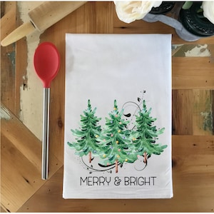 Peut inclure: Serviette de cuisine blanche avec une illustration aquarelle de trois sapins de No&euml;l avec des lumi&egrave;res et le texte "Merry & Bright".