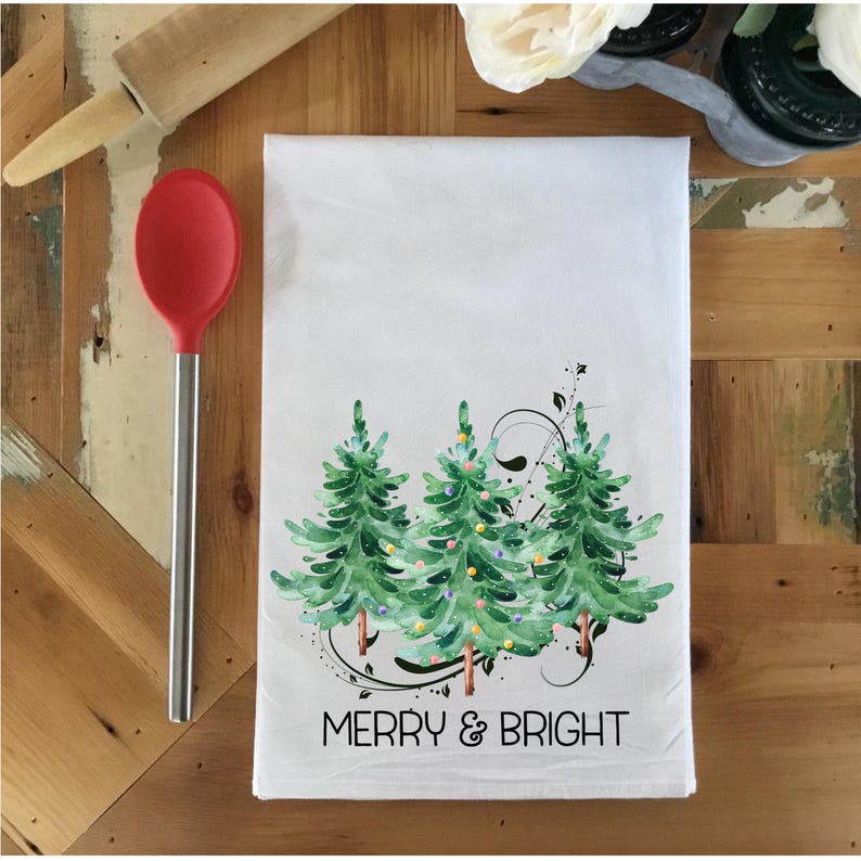 Peut inclure: Serviette de cuisine blanche avec une illustration aquarelle de trois sapins de No&euml;l avec des lumi&egrave;res et le texte "Merry & Bright".