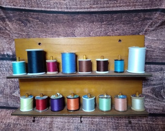 May include: A wooden spool rack with two shelves holds a variety of colorful thread spools. The spools are arranged in rows and are made of different materials, including plastic, metal, and wood.