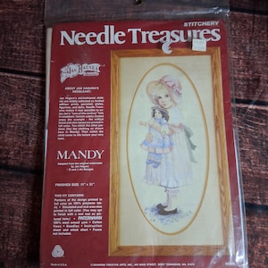 May include: A Needle Treasures stitchery kit titled Mandy, featuring a printed illustration of a child holding a doll. The packaging is red with white text, displaying a finished size of 11 inches by 21 inches and listing kit contents.