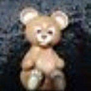 May include: A small, brown, plush teddy bear sitting with its legs crossed. The bear has a light brown nose and black eyes.