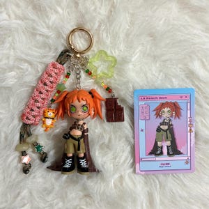 May include: A keychain featuring an orange-haired character figurine, a braided pink cord, a small orange cat charm, a green star, and a brown chocolate bar charm. Beside it is a rectangular card with the text Lil Peach Riot and Gigi BM.
