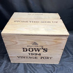 May include: Wooden crate with the text "DOW'S 1994 VINTAGE PORT" printed on the front. The top of the crate reads "STORE THIS SIDE UP" and "WEIGHT 21 KG". The crate is made of light-colored wood.