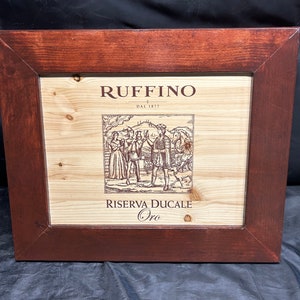 May include: A framed wooden sign featuring the Ruffino logo and a detailed illustration of people and a dog. The frame is a dark brown color, and the sign's background is light-colored wood. The text "Riserva Ducale Oro" is visible.