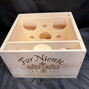 Op de afbeelding: Lichtgekleurde houten wijnkist met cirkelvormige uitsparingen voor flessen. De voorkant van de kist heeft de tekst "Far Niente" boven een illustratie van een gebouw en de tekst "2022 Cabernet Sauvignon - Napa Valley."