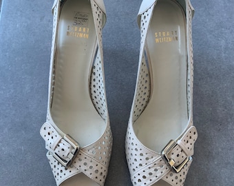 Puede incluir: Un par de tacones blancos de cuero con puntera abierta y detalle de hebilla. Los tacones tienen un diseño perforado y el nombre de la marca "Stuart Weitzman" está impreso en el interior de cada zapato.