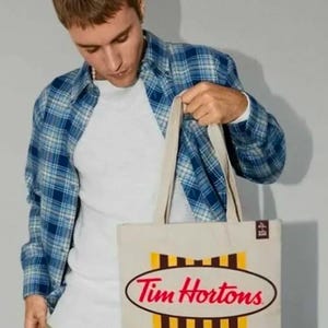Puede incluir: Una bolsa de lona blanca con un diseño de rayas marrones y amarillas. La bolsa presenta dos logotipos ovalados, uno para Tim Hortons y otro para Tim Biebs.