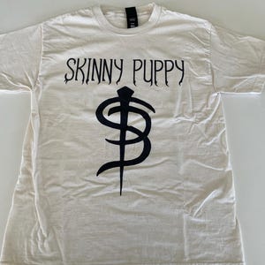 May include: A short-sleeved, off-white crew neck t-shirt featuring the text SKINNY PUPPY printed in black across the chest, positioned above a large, stylised black symbol resembling a stylised dollar sign with a vertical line through the centre.