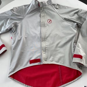 Pode incluir: Uma jaqueta prateada com detalhes vermelhos e um logotipo de raio. A jaqueta tem fecho de zíper e gola alta.