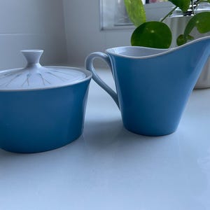 Op de afbeelding: Een lichtblauwe keramische suikerpot met bijpassend deksel en een lichtblauwe melkkan. Het deksel van de suikerpot heeft een witte bovenkant met een stralend patroon van dunne gouden en blauwgroene lijnen. Beide stukken hebben een gladde blauwe afwerking.