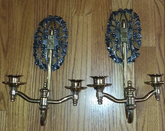 May include: A pair of antique brass wall sconces with ornate black metal accents. Each sconce has three candle holders and a decorative floral design.