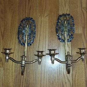May include: A pair of antique brass wall sconces with ornate black metal accents. Each sconce has three candle holders and a decorative floral design.