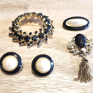 May include: A collection of vintage jewelry pieces, including a circular brooch with black stones and gold-tone metal, a black and gold oval pin, a brooch with a black stone and tassel, and a pair of black and gold earrings with white centers.