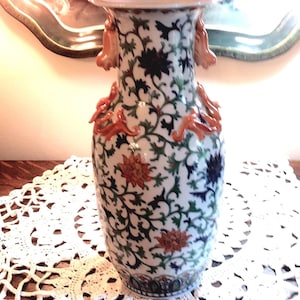May include: A tall porcelain vase featuring a white base with an intricate pattern of green vines and multi-colored floral motifs in orange and dark blue. The neck of the vase is adorned with sculpted orange animal-shaped handles on both sides.