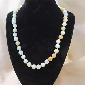 May include: A necklace made of light green, white, and beige round beads. The necklace is displayed on a black mannequin.