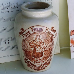 May include: A cream-coloured ceramic jar with a brown transfer print featuring a person holding a bowl and the text PURE FRESH CREAM FROM THE WIGTOWNSHIRE CREAMERY CO STRANRAER. Beside it is a vintage postcard showing a child with a mirror and dog.