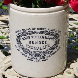May include: A vintage cream-coloured ceramic jar overflowing with pink and red flowers. The jar features a black and white label for "JAMES KEILLER & SONS DUNDEE MARMALADE". The flowers are a mix of shades, creating a colourful display.