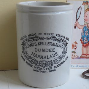 May include: A cream-coloured ceramic Dundee Marmalade jar with black text and a decorative emblem. The jar is cylindrical. A vintage postcard and antique keys are also in the image. The jar has a diameter of approximately 10 cm.