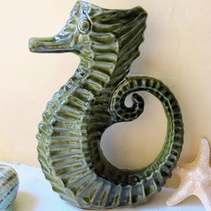 May include: A green ceramic seahorse figurine with a curled tail, a long snout, and textured details. A white starfish and a blue and white seashell are next to the seahorse. The figurine is approximately 25 cm tall.