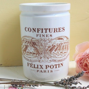 May include: White ceramic jar with a brown label that reads "Confitures Fines Felix Potin Paris" and a vintage illustration of a factory. The jar is used for storing preserves.