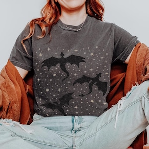 May include: Dark grey t-shirt with a black dragon design and a starry pattern. The shirt is paired with light blue jeans, white trainers, and a brown jacket. The image has a casual and relaxed feel.