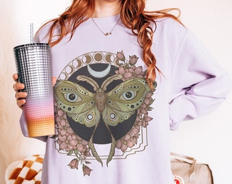 May include: A lavender sweatshirt with a graphic print of a moth with large eyes, surrounded by flowers and a crescent moon. The moon is surrounded by three circles.