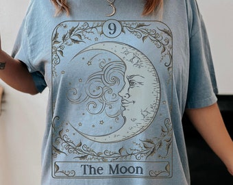 May include: A light blue short-sleeve t-shirt featuring a graphic print of a tarot card design. The print depicts a crescent moon with a face, surrounded by stars and swirling clouds, enclosed in a rectangular border with the text The Moon.